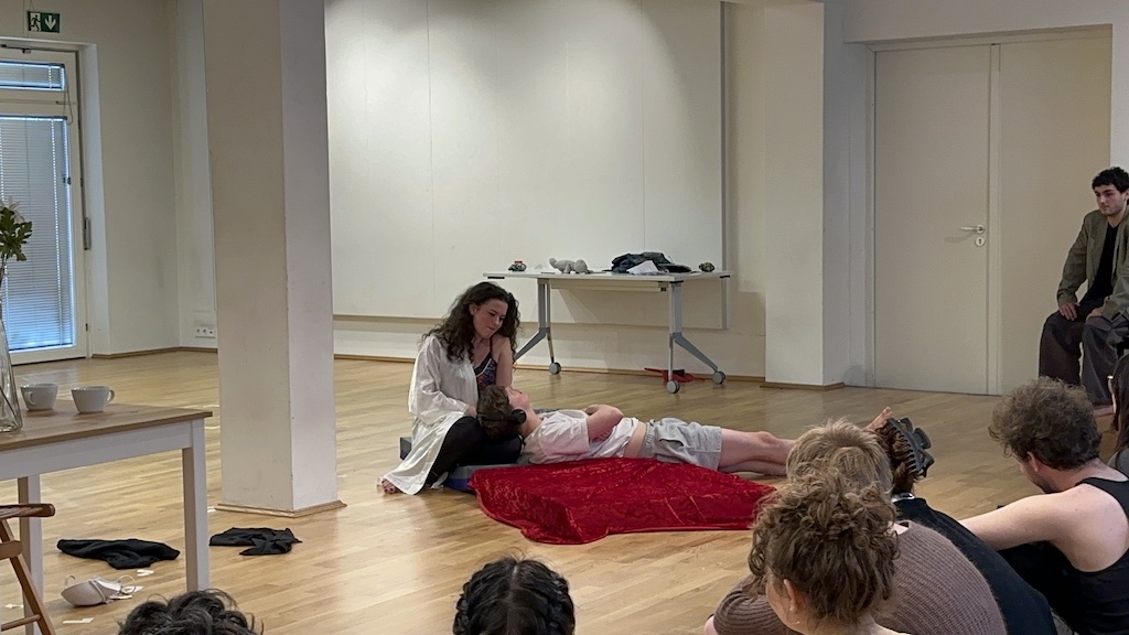 An actor lays prone on a red blanket as a second actor sits near them, cradling their head in their lap in mid-performance of 'Faith Hope Charity'. A third actor looks on from the right side of the photo as a few students in the audience watch the performance.